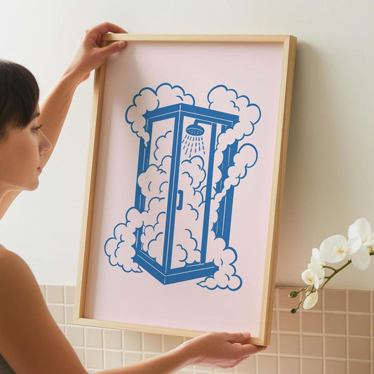 A woman holds a framed print of a blue shower stall surrounded by pink clouds on a light pink background.