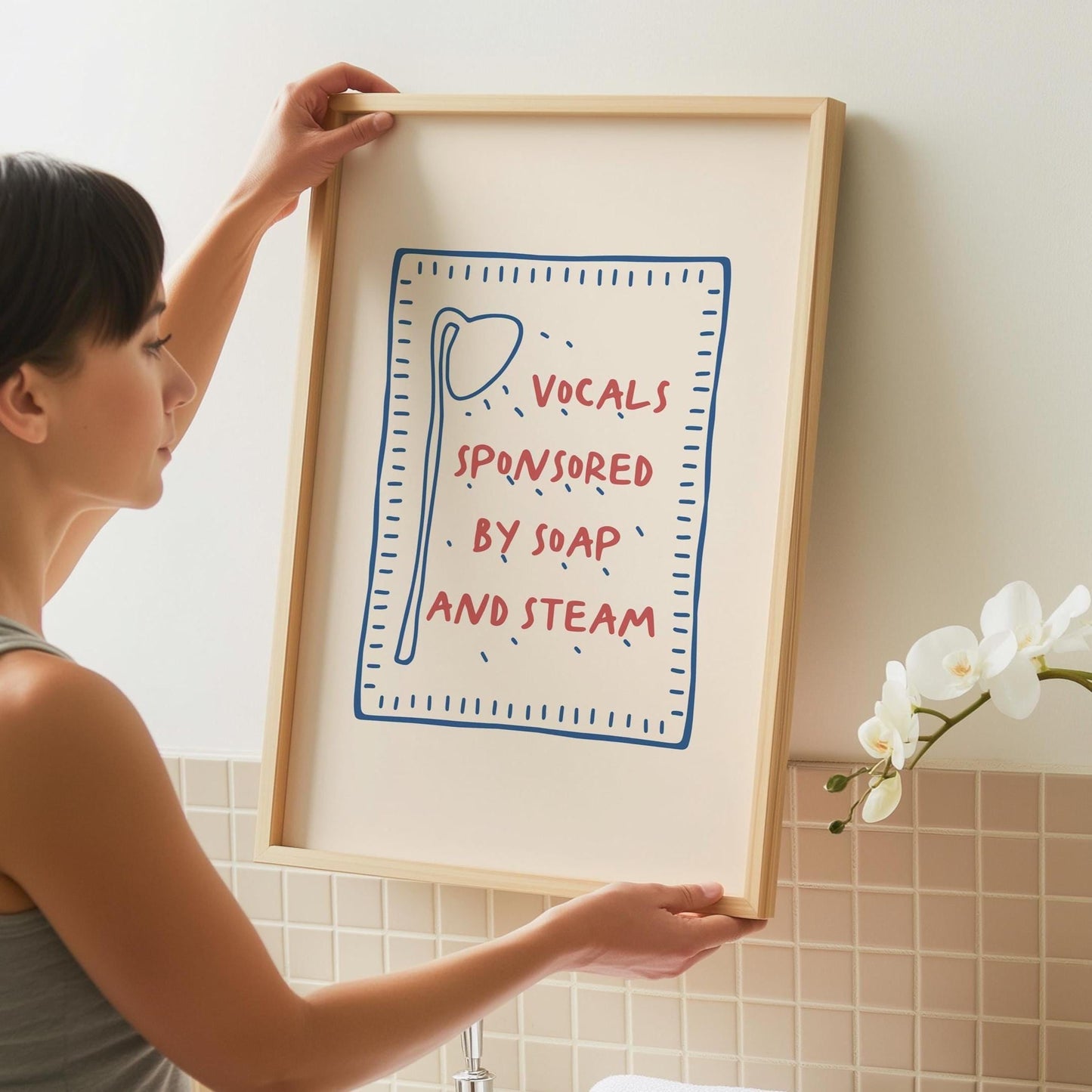 A person holds a framed print with a light wood frame, featuring a red and blue design on a cream background that reads "VOCALS SPONSORED BY SOAP AND STEAM."