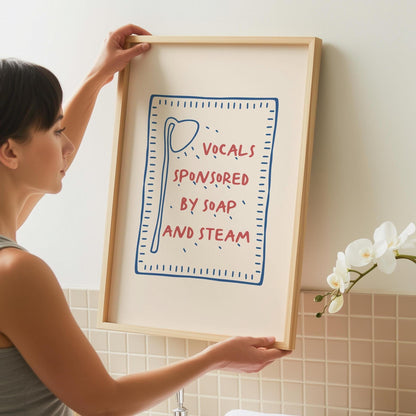 A person holds a framed print with a light wood frame, featuring a red and blue design on a cream background that reads "VOCALS SPONSORED BY SOAP AND STEAM."