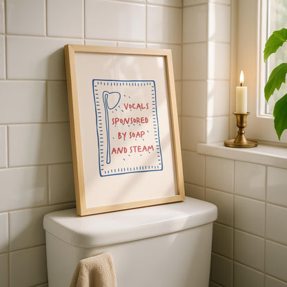 A framed print with a light wood frame leans against a white tiled wall on top of a toilet, displaying a red and blue design on a cream background that reads "VOCALS SPONSORED BY SOAP AND STEAM."