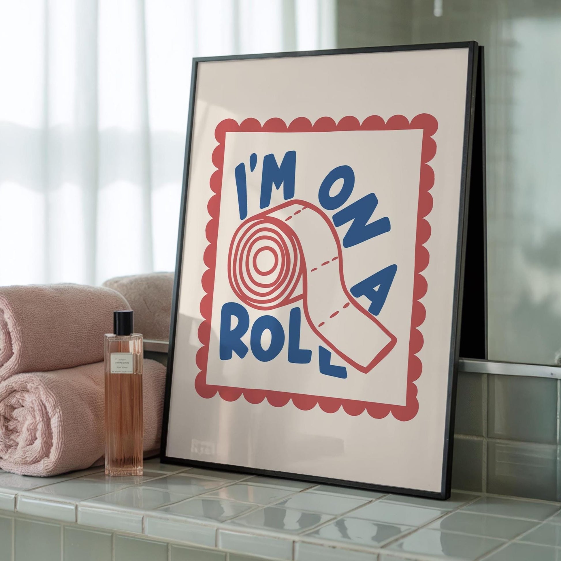 A black-framed print with "I'M ON A ROLL" in blue text and a red toilet paper roll on a pink scalloped border, leaning against a mirror next to stacked pink towels and a perfume bottle.