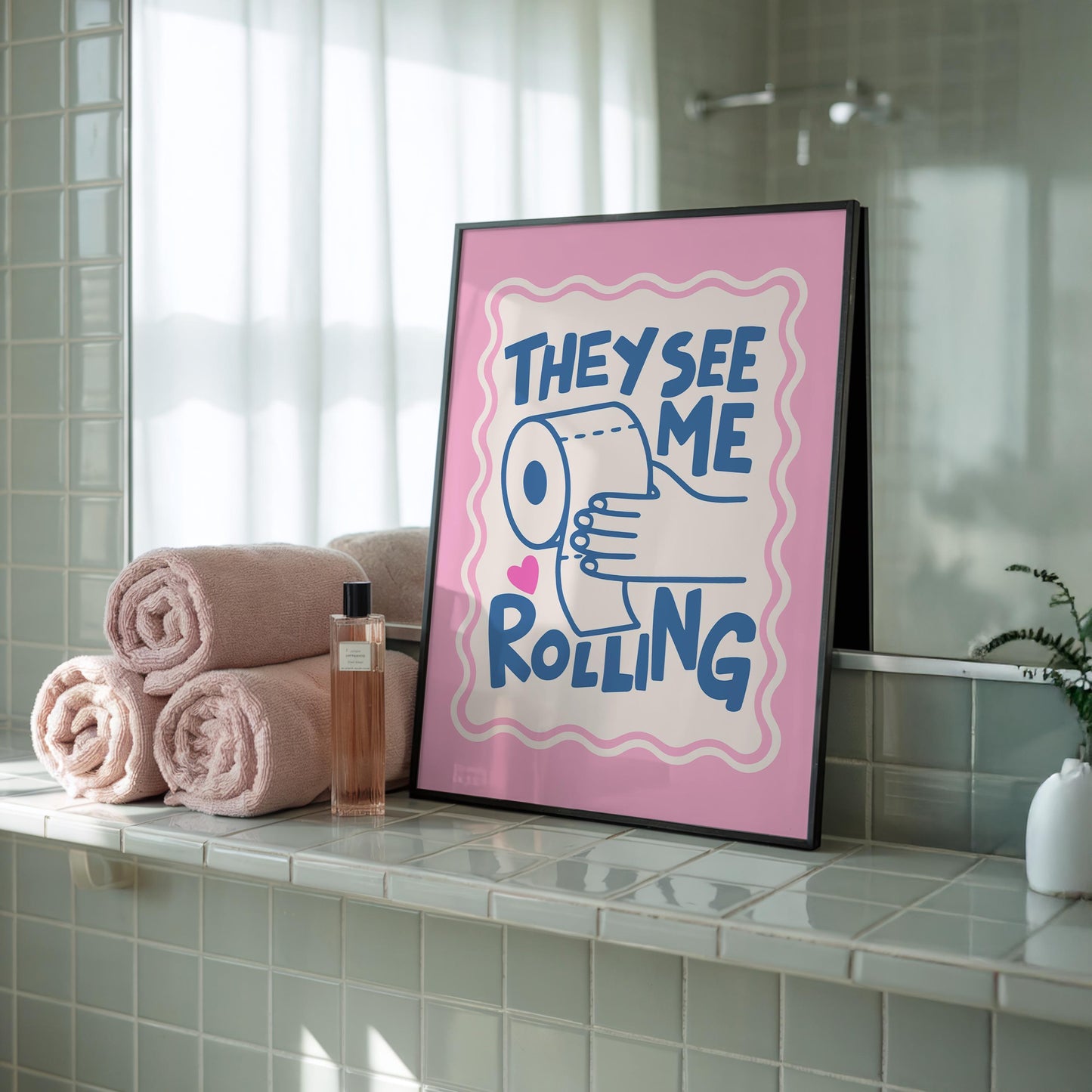 A framed print with a pink border, a light pink background, and the words "THEY SEE ME ROLLING" in blue, with a hand holding a toilet paper roll, sits on a green tiled bathroom counter next to stacked pink towels.