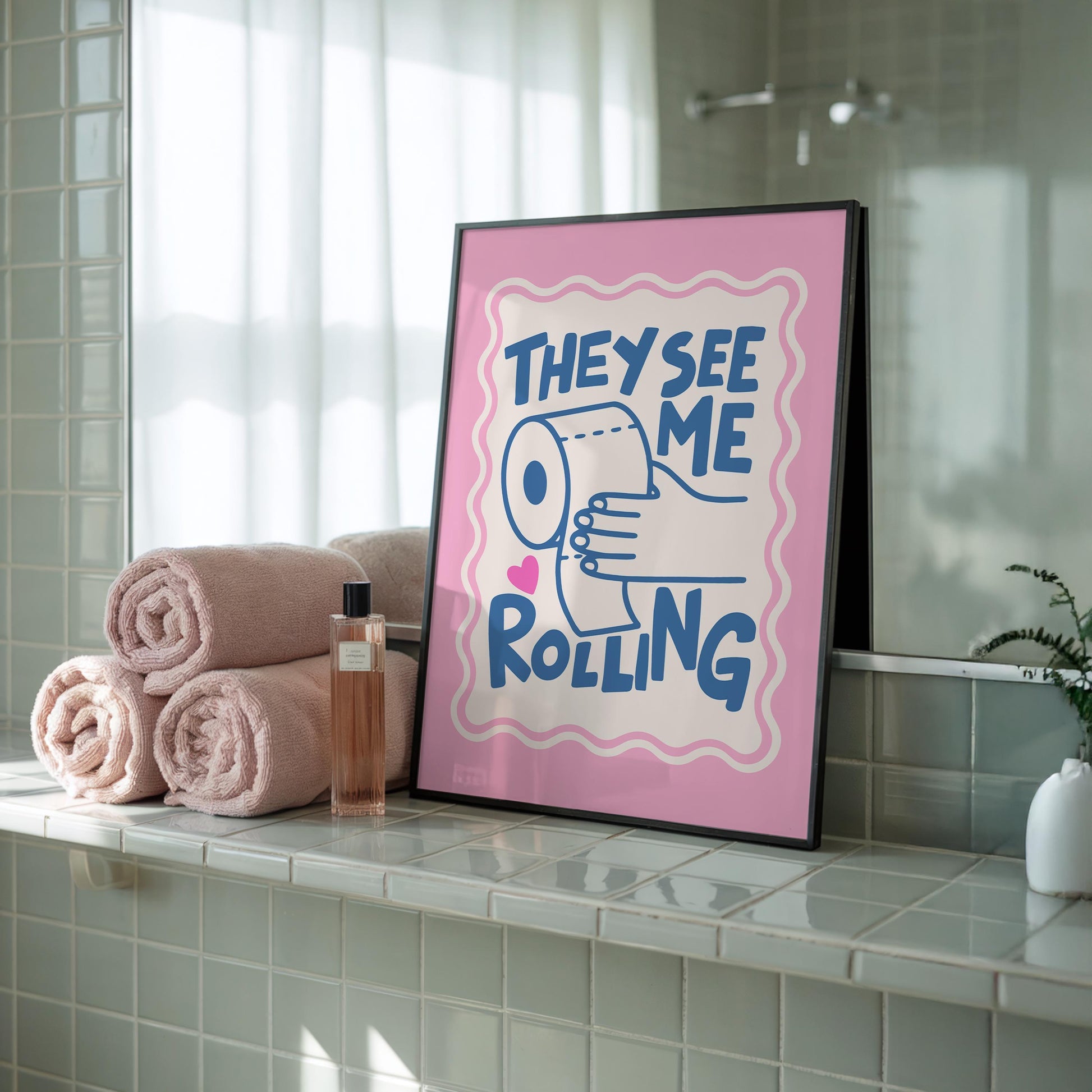 A framed print with a pink border, a light pink background, and the words "THEY SEE ME ROLLING" in blue, with a hand holding a toilet paper roll, sits on a green tiled bathroom counter next to stacked pink towels.