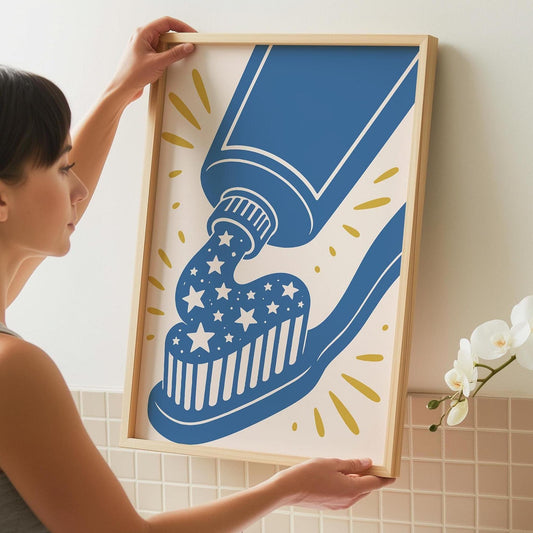 A woman holds up a framed print of a blue toothpaste tube squeezing white, star-shaped toothpaste onto a blue toothbrush, all against a cream background with yellow starbursts.