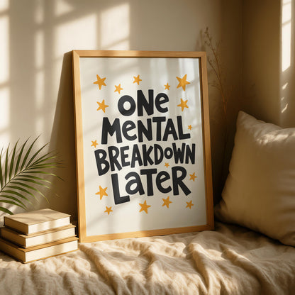 A light wood-framed print with "ONE MENTAL BREAKDOWN LATER" in black text and yellow stars on a cream background, leaning against a wall on a textured beige blanket with a stack of books and a plant.