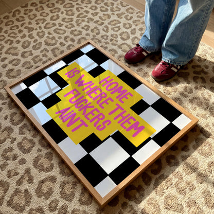 A framed print with a black and white checkerboard background and a yellow cross with the pink text "HOME IS WHERE THEM FUCKERS AINT" lies on a leopard print rug, with a person's feet in red shoes visible.