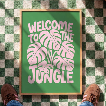 A framed green print with pink monstera leaves and the text "Welcome to the Jungle" sits on a green and white checkered floor, with a person's brown shoes visible at the bottom.