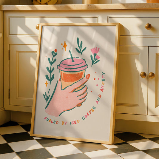 A framed print on a kitchen counter shows a hand holding an iced coffee with the text "FUELED BY ICED COFFEE AND ANXIETY" surrounded by green leaves and pink flowers.