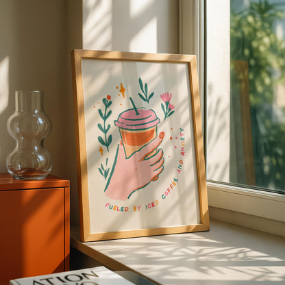 A framed print on a sunlit windowsill shows a hand holding an iced coffee with the text "FUELED BY ICED COFFEE AND ANXIETY" surrounded by green leaves and pink flowers.