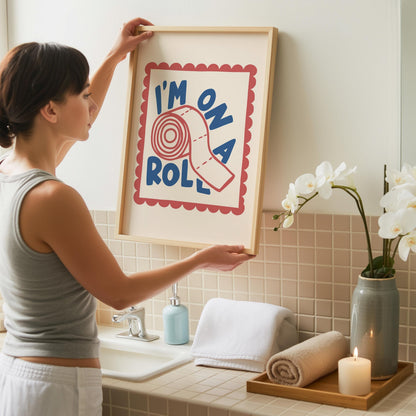 A woman in a gray tank top hangs a light wood-framed print with "I'M ON A ROLL" and a toilet paper roll, above a white sink with a blue soap dispenser and white towels.
