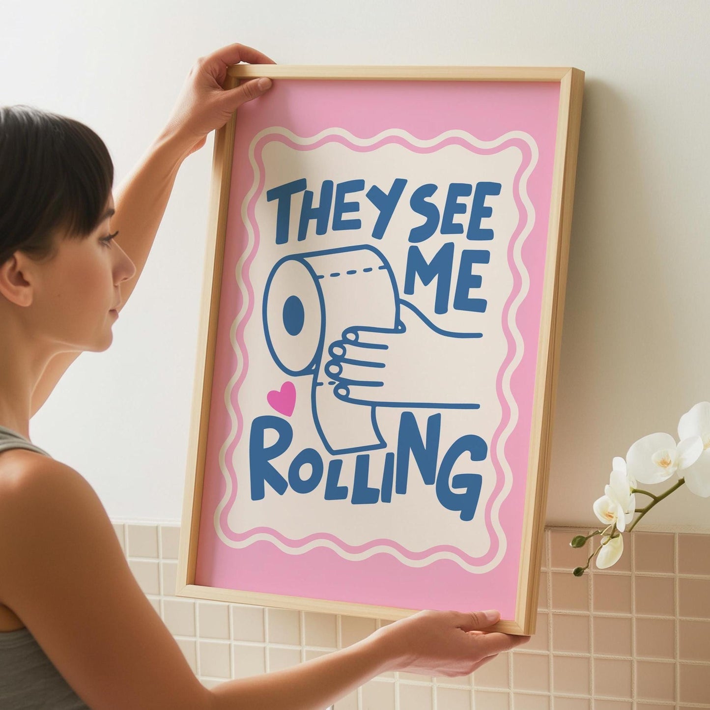 A person holds up a framed print with a pink border, a light pink background, and the words "THEY SEE ME ROLLING" in blue, with a hand holding a toilet paper roll.